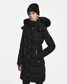 A woman wears the Rudsak KEHLANI - L124540 Black knee-length quilted down coat with a faux fur-trimmed hood and a black knit beanie, standing against a plain white background.