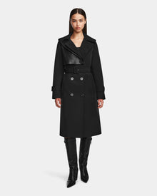 A woman stands facing forward, wearing the Rudsak KINDA - 8124690 Black trench coat with leather detailing and double-breasted buttons, paired with knee-high black boots, against a plain white background.