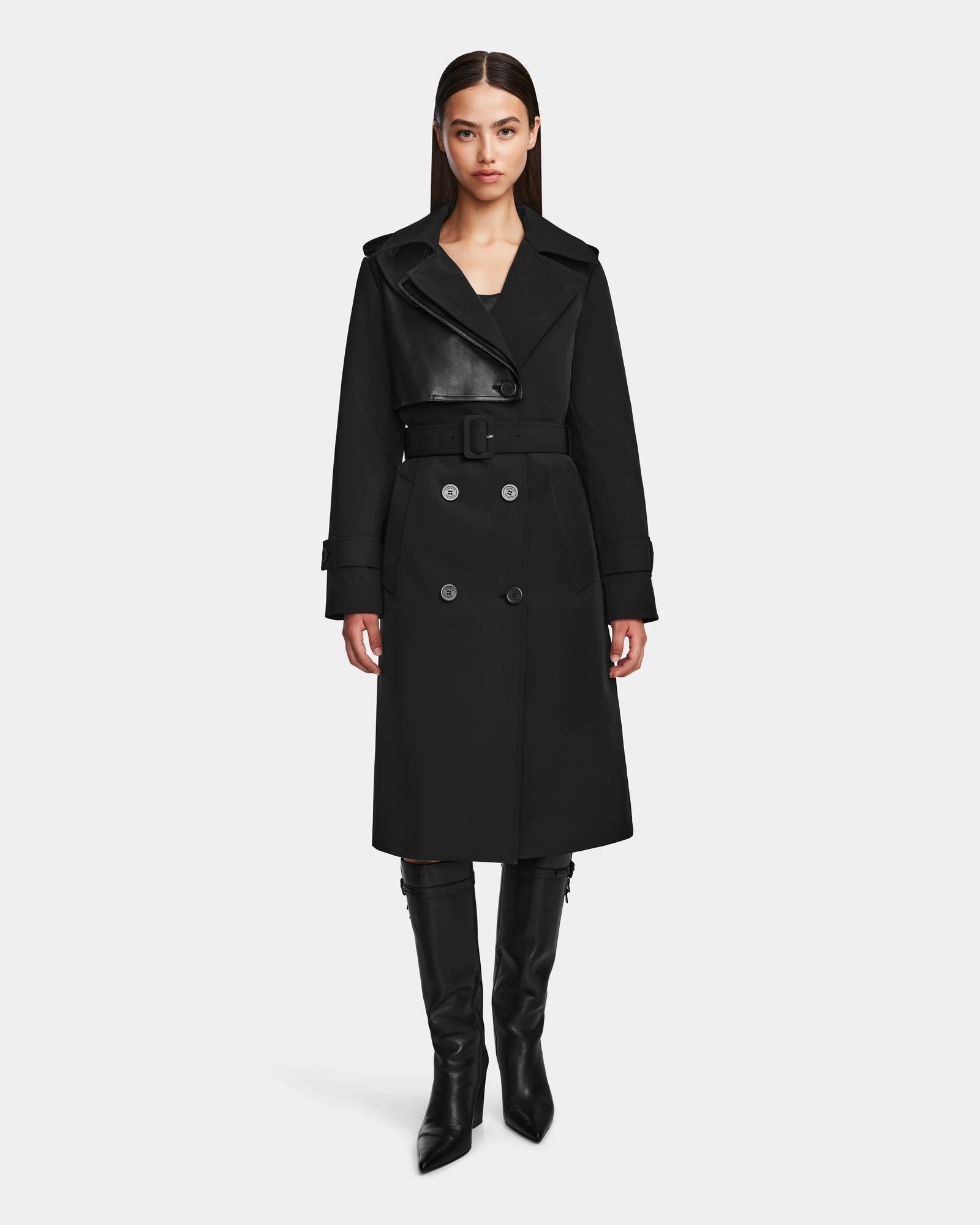 A woman stands facing forward, wearing the Rudsak KINDA - 8124690 Black trench coat with leather detailing and double-breasted buttons, paired with knee-high black boots, against a plain white background.