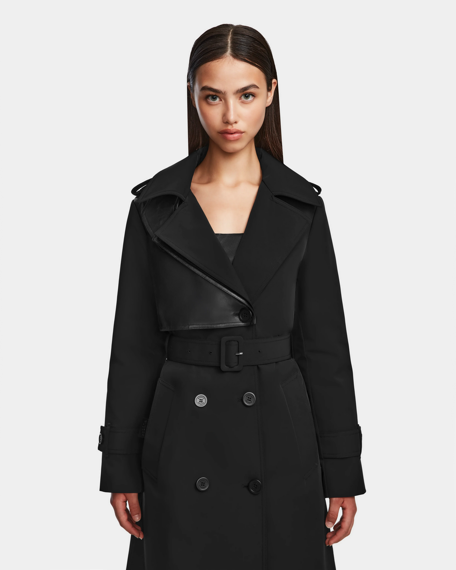 A woman with straight dark hair wears the Rudsak KINDA - 8124690 Black trench coat, featuring leather details, a belted waist, and double-breasted buttons, standing against a plain white background.