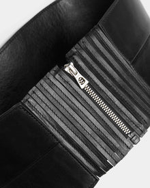 Close-up of the Rudsak LEA - 8424519 Black wide waist belt in black leather with ribbed detailing and a shiny silver zipper, partially unzipped, displayed against a light gray background—a versatile accessory.