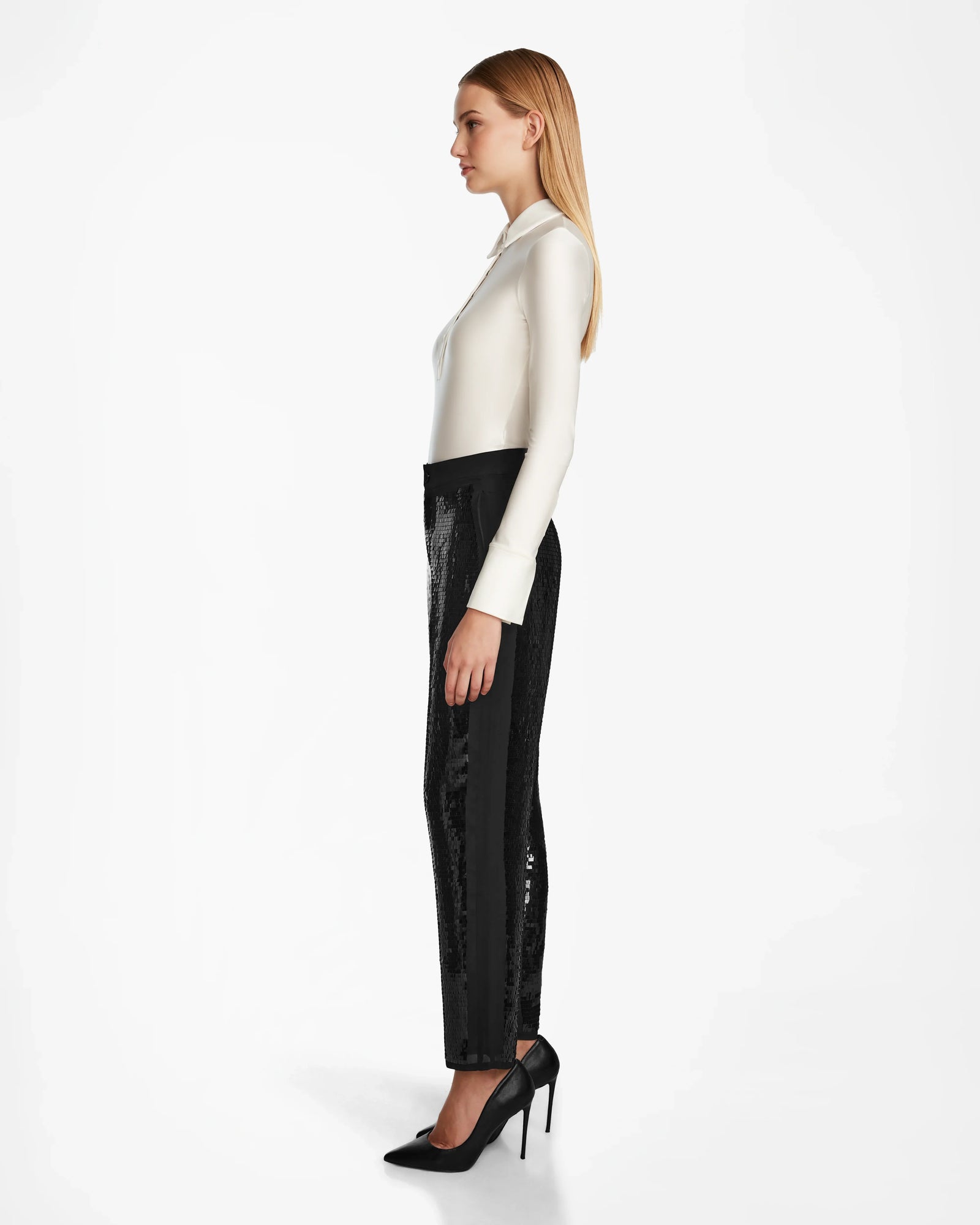 A woman with straight blonde hair stands in profile, wearing a long-sleeve white blouse, women’s sequin pants, and black high-heeled Rudsak LEAR - 8524553 shoes against a plain white background.
