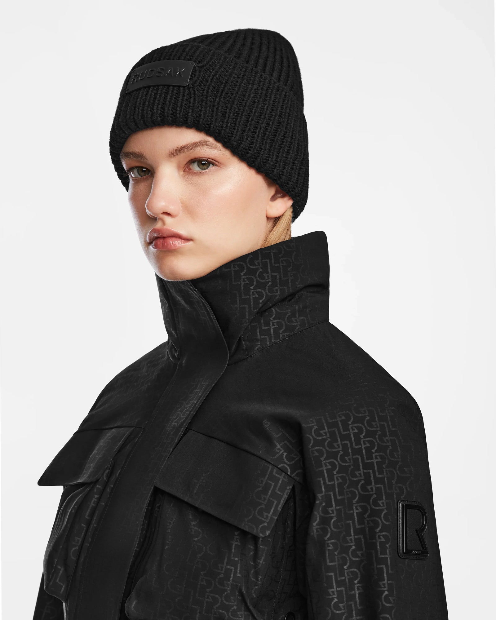 A person wearing a textured black beanie and the Rudsak LUX MO - 8124570 Monogram Print Black ski jacket looks forward with a neutral expression against a plain light background.