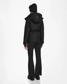 A person wearing the Rudsak LUX MO - 8124570 Monogram Print Black women’s ski jacket with RECCO rescue tech faces away, paired with black pants, boots, and a beanie, displaying the back of the outfit against a plain white background.