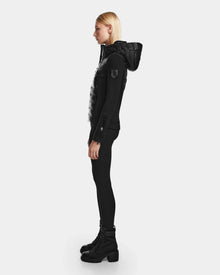 A person with straight blonde hair stands in profile, wearing an all-black outfit featuring the RUDSAK MABS - 8122020 BLACK puffer jacket, fitted pants, and chunky lace-up boots against a plain light background.