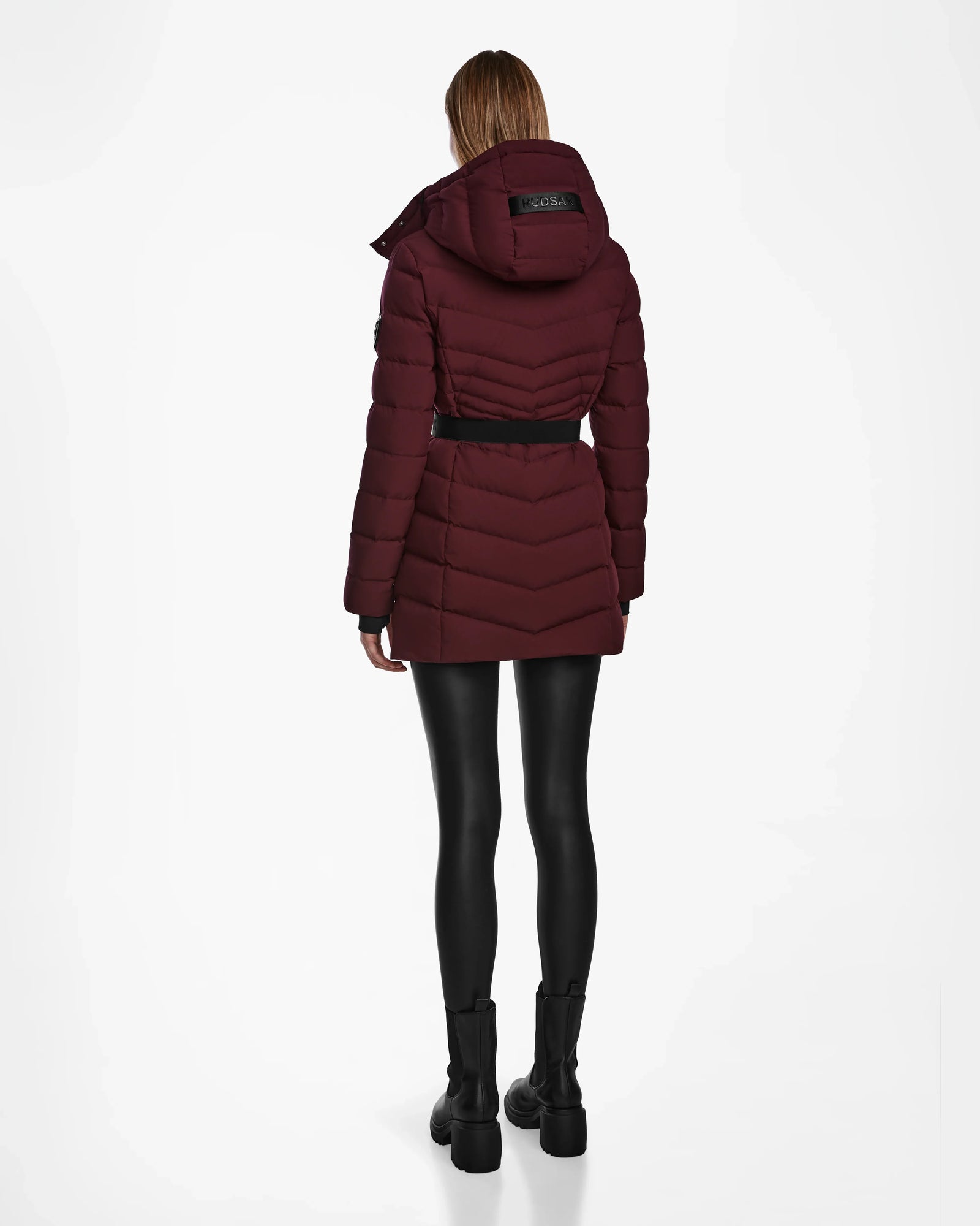 A person stands facing away, wearing the Rudsak MALAYA M - L123554 Wine womens down puffer jacket with herringbone quilting and hood, styled with a black belt, leggings, and boots against a plain white background.