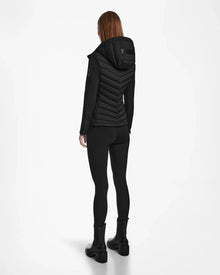 A person with long hair faces away, wearing the Rudsak MALYA M - L123547 Black down quilted jacket, black leggings, and black boots against a plain white background.