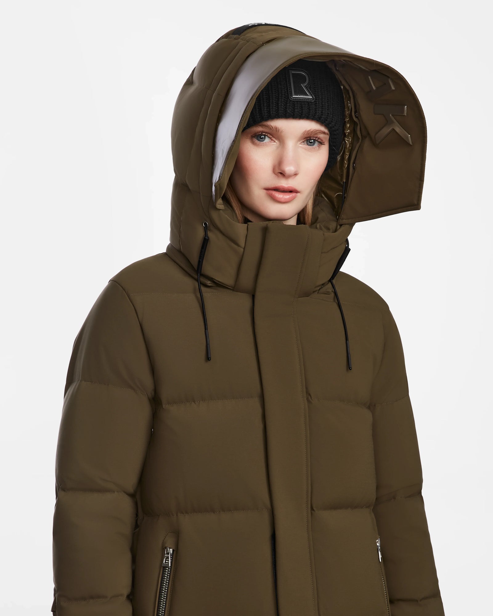 A woman wears the Rudsak MAO M - 8123598 Army olive green down puffer coat with hood up and a black knit beanie, made from weatherproof recycled Stormshell, standing against a plain white background.