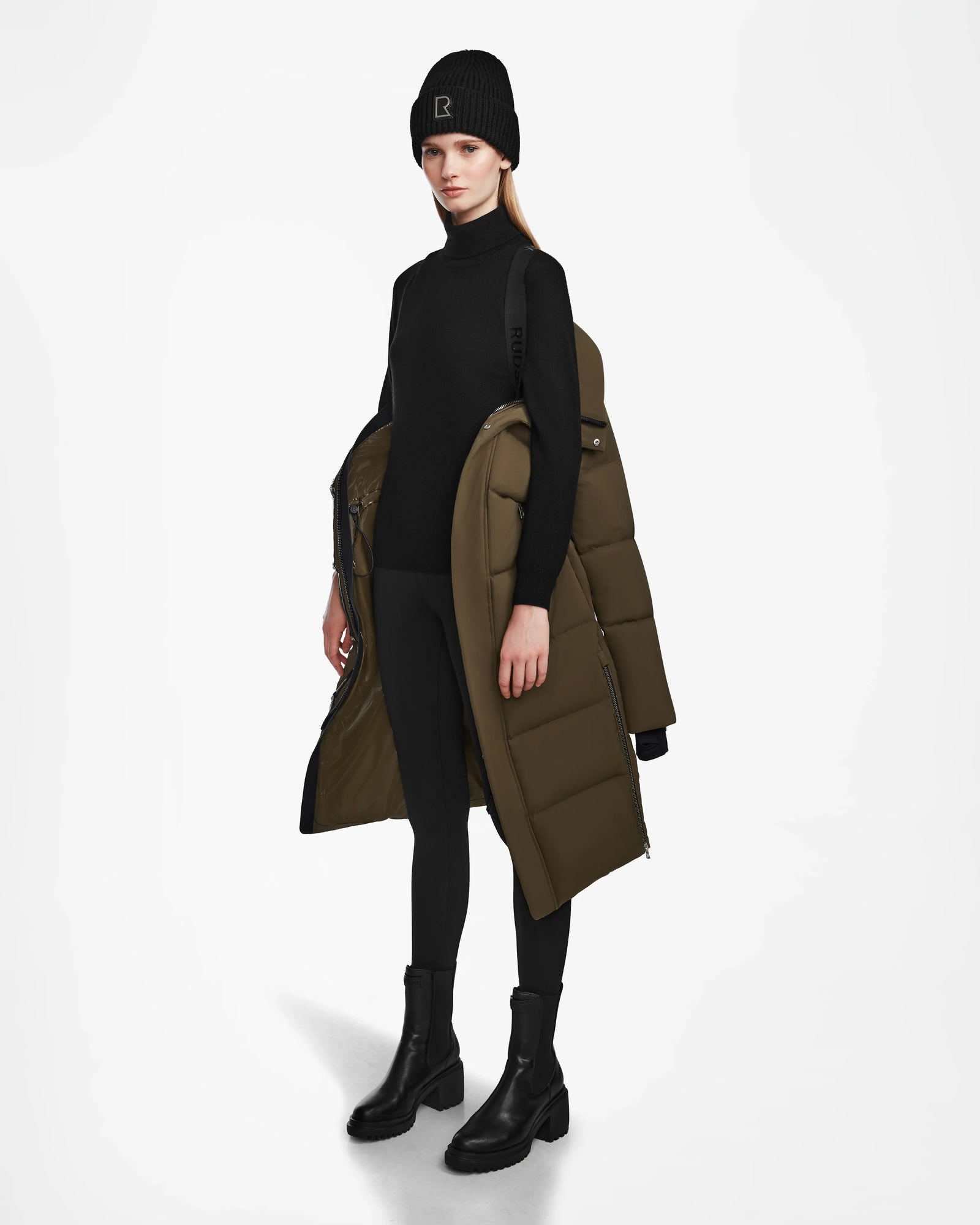 A woman stands against a white background in a sleek black outfit, holding the Rudsak MAO M - 8123598 Army womens down puffer coat with weatherproof recycled Stormshell draped over her shoulders.