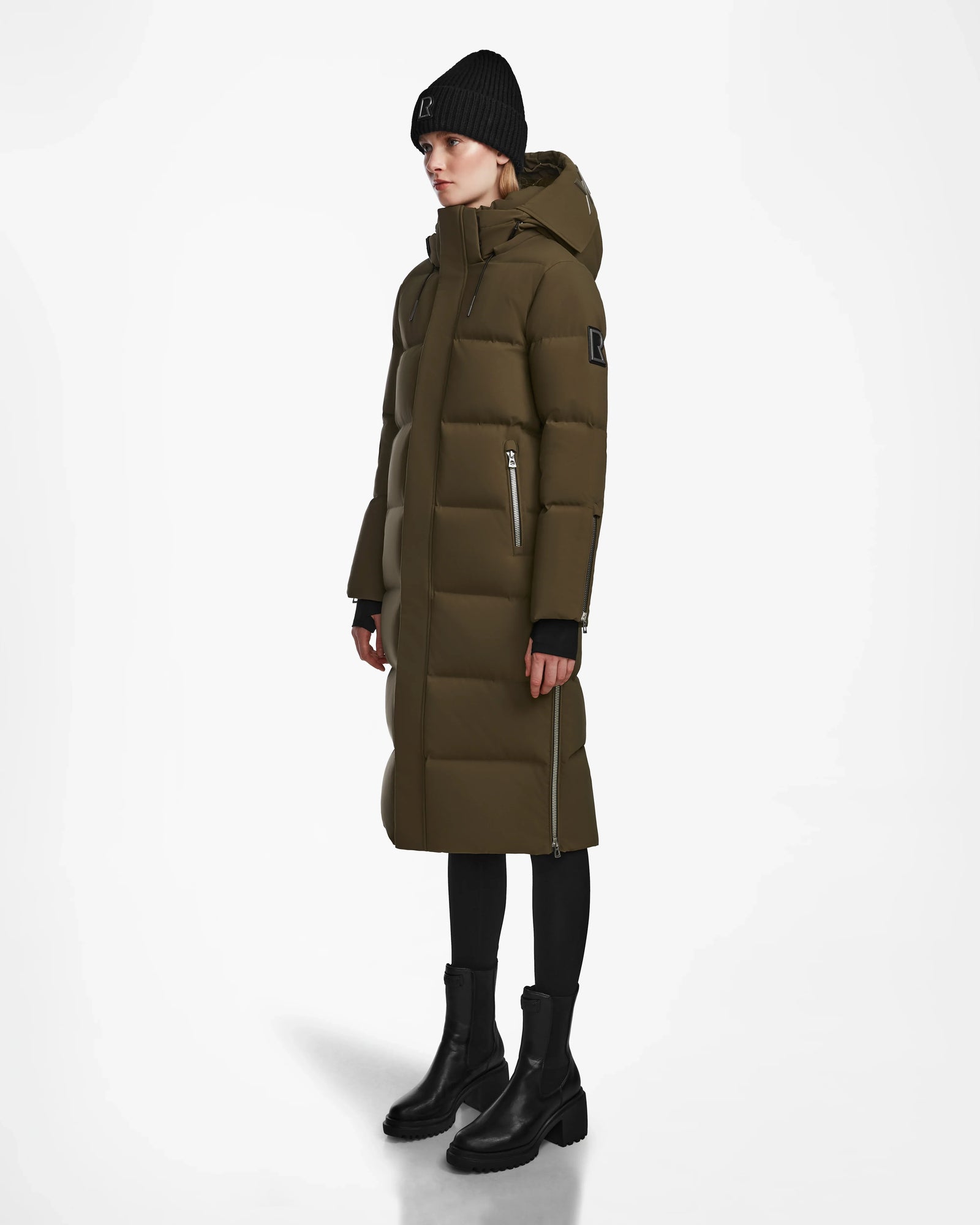 A person wearing the Rudsak MAO M - 8123598 Army women’s down puffer coat with a hood, black beanie, leggings, and ankle boots stands against a plain white background.