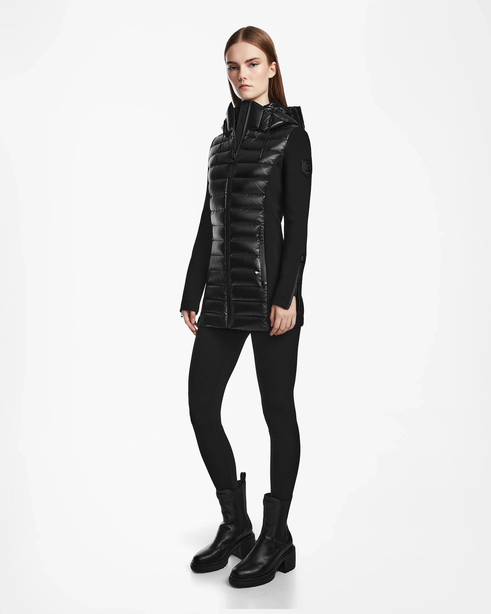 A woman with straight brown hair and a neutral expression stands against a white background wearing the Rudsak MARRY - L121003 BLACK puffer jacket over a black top, black leggings, and boots.