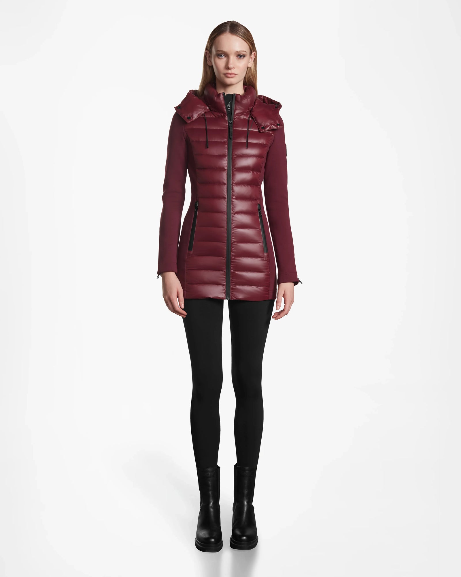 A woman faces forward, wearing the Rudsak MARRY - L121003 Wine long puffer with hood, black leggings, and black ankle boots against a plain white background.