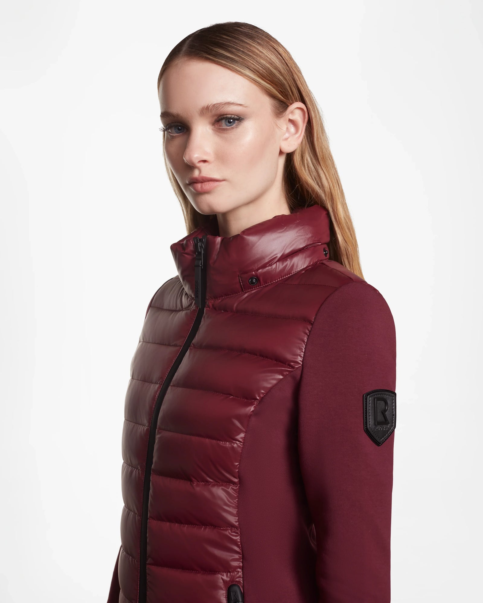A woman with straight blond hair models the Rudsak MARRY - L121003 Wine women’s puffer jacket, featuring a high collar and black sleeve patch, against a white background while looking slightly to the side.