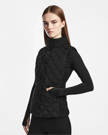 A woman with straight brown hair wears the Rudsak MATILDA - L124504 Black womens light vest with diamond quilting over a black long-sleeve top, standing against a plain white background, one hand in her pocket.