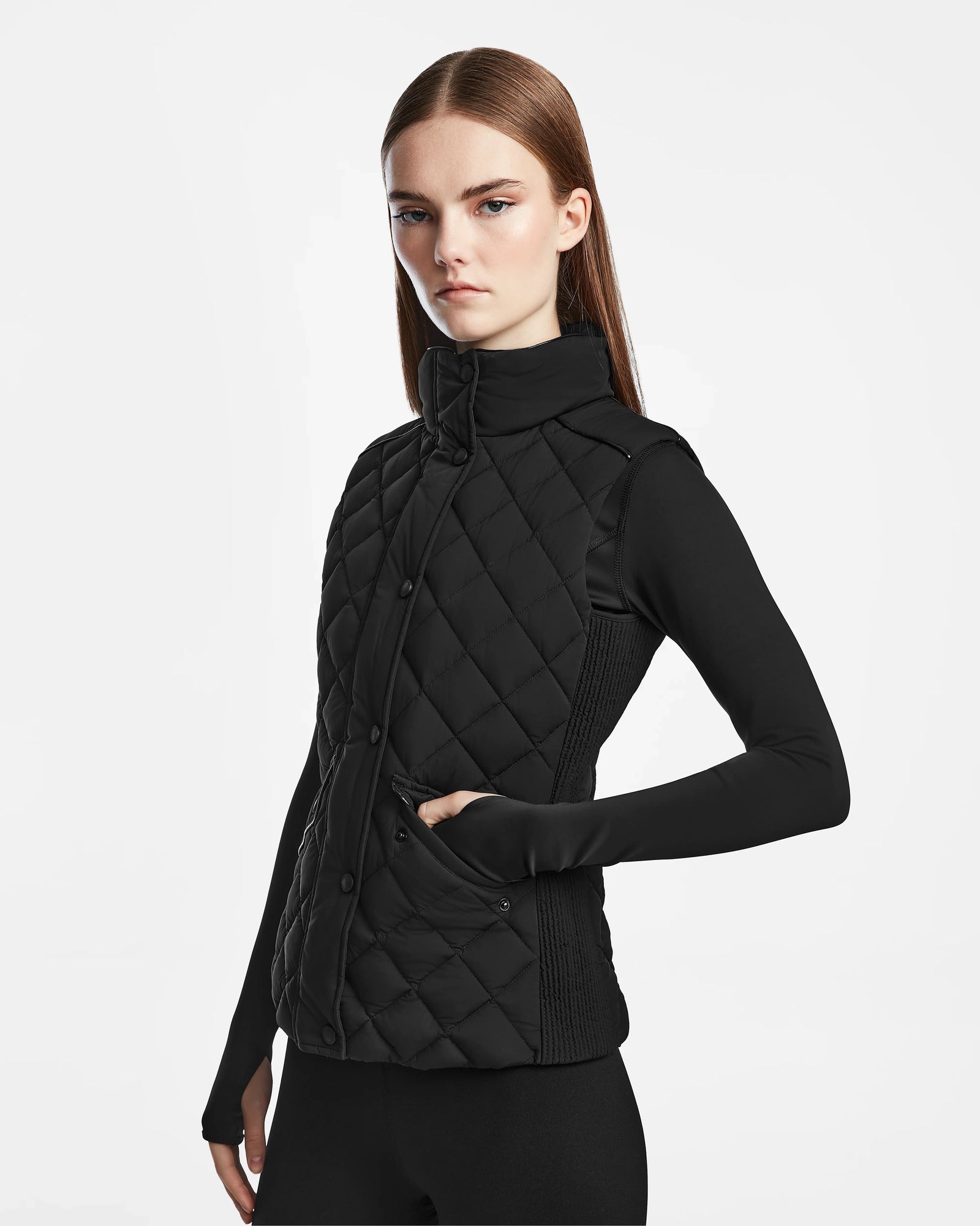 A woman with straight brown hair wears the Rudsak MATILDA - L124504 Black womens light vest with diamond quilting over a black long-sleeve top, standing against a plain white background, one hand in her pocket.