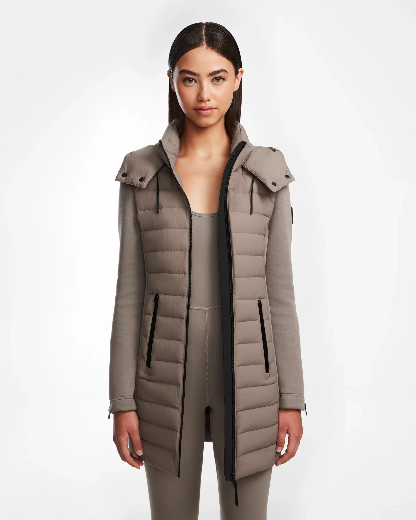 A woman with straight dark hair wears the Rudsak MERINA M - 8124030 Cement, a taupe long quilted down jacket with hood and black zipper, standing against a plain light background, facing forward with a neutral expression.