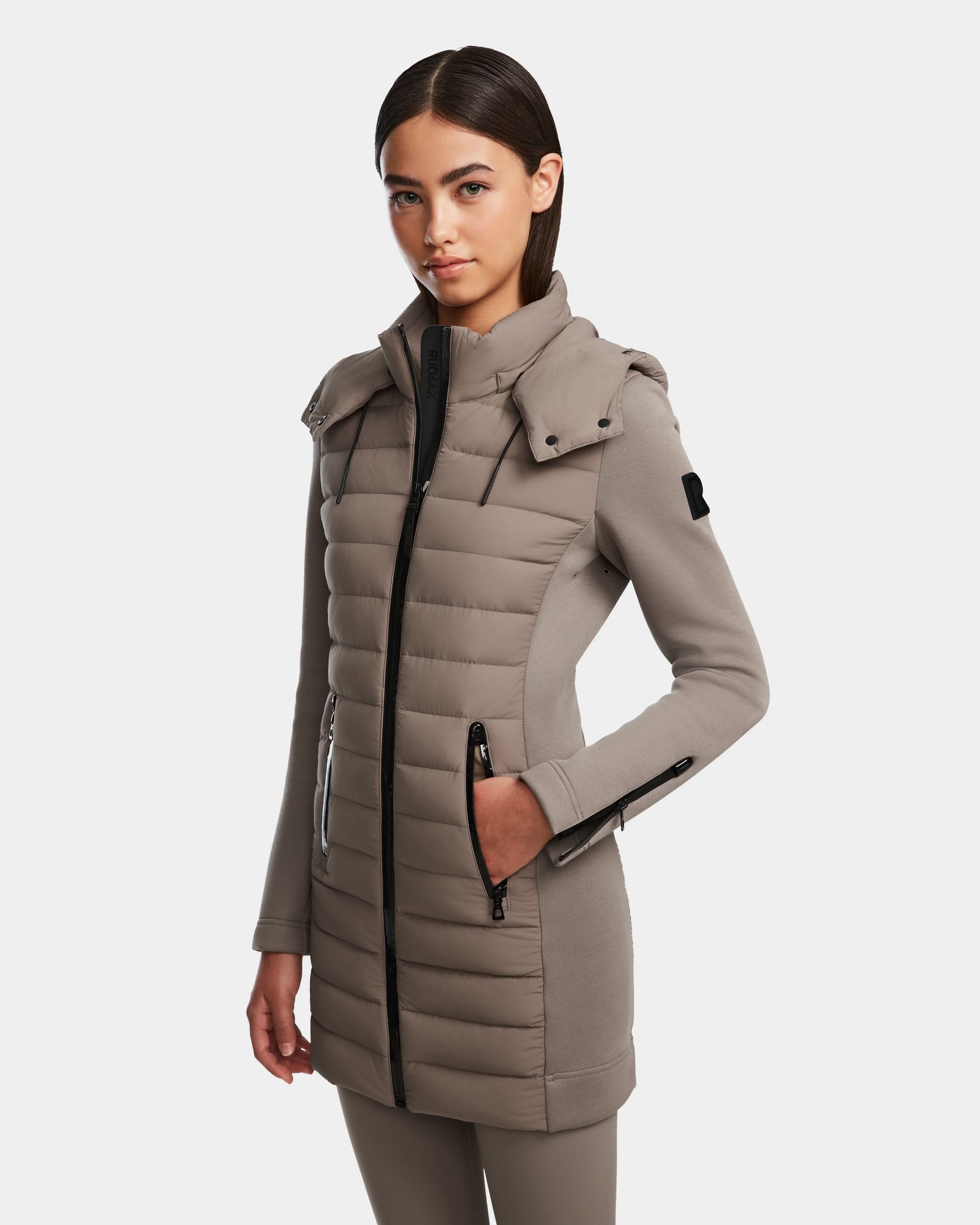A woman with straight dark hair wears the Rudsak MERINA M - 8124030 Cement, a long light taupe quilted down jacket with a zip front, high collar, and side pockets. She stands against a white background, looking confidently at the camera.