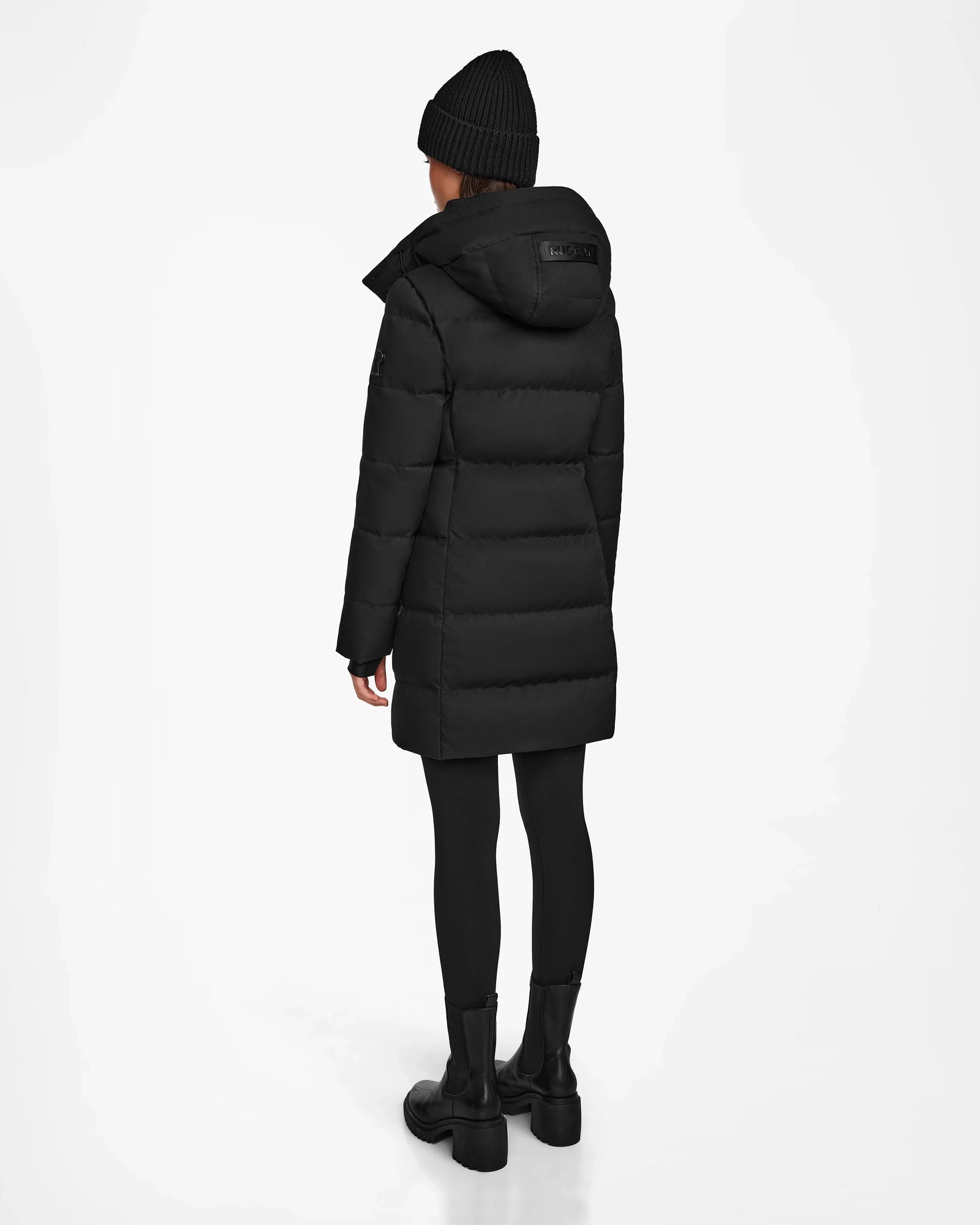 A person wears the Rudsak MIRIAM - L122523 BLACK womens down puffer coat with black pants, boots, and a knit beanie, standing against a white background—a chic winter staple.
