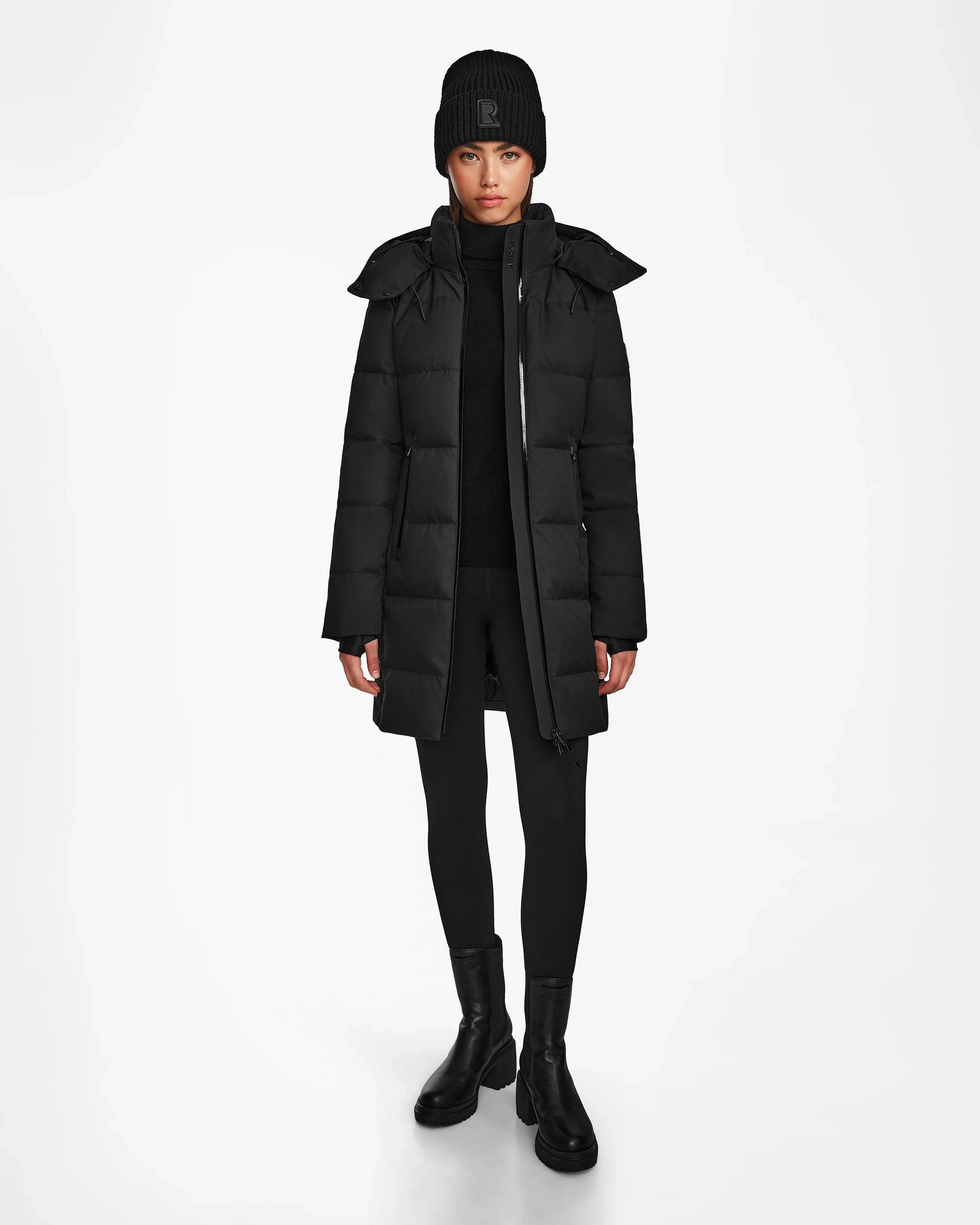 A person faces forward wearing the Rudsak MIRIAM - L122523 BLACK womens down puffer coat, paired with a black knit beanie, black pants, and black boots against a plain white background.