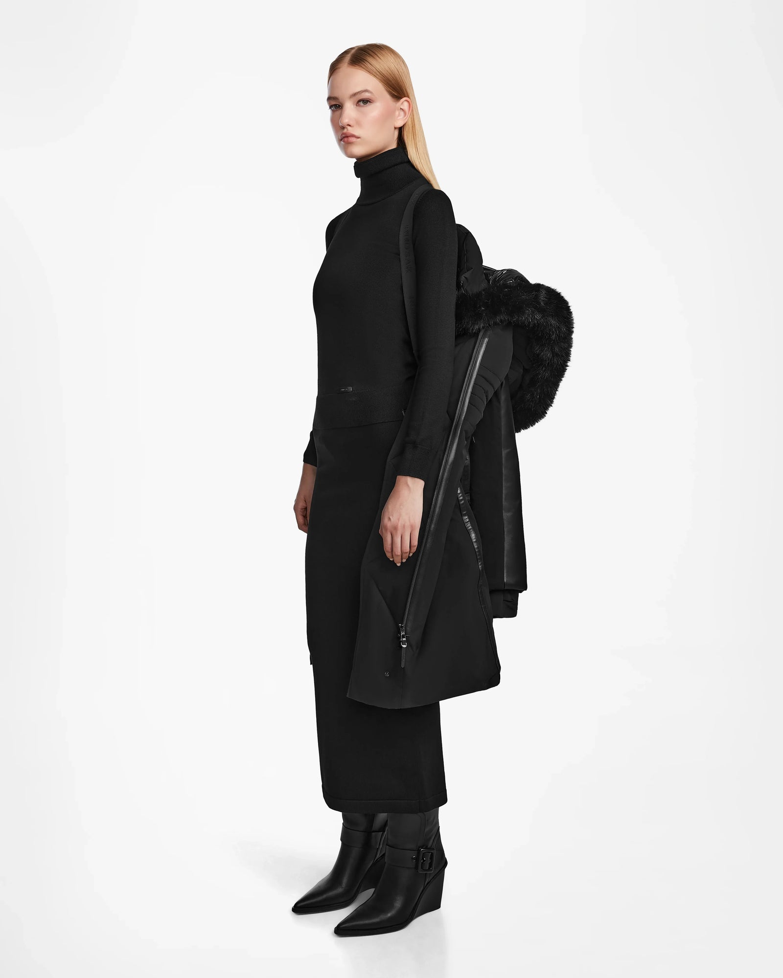 A woman with straight blonde hair stands against a white background, wearing a black turtleneck, skirt, and ankle boots. She holds the Rudsak MISALA - L124512 Black women’s long down coat over her arm.