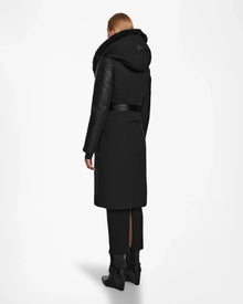 A person with straight, light brown hair is shown from the back wearing the Rudsak MISALA - L124512 Black womens long down coat with a hood, black pants, and black heeled boots against a plain white background.