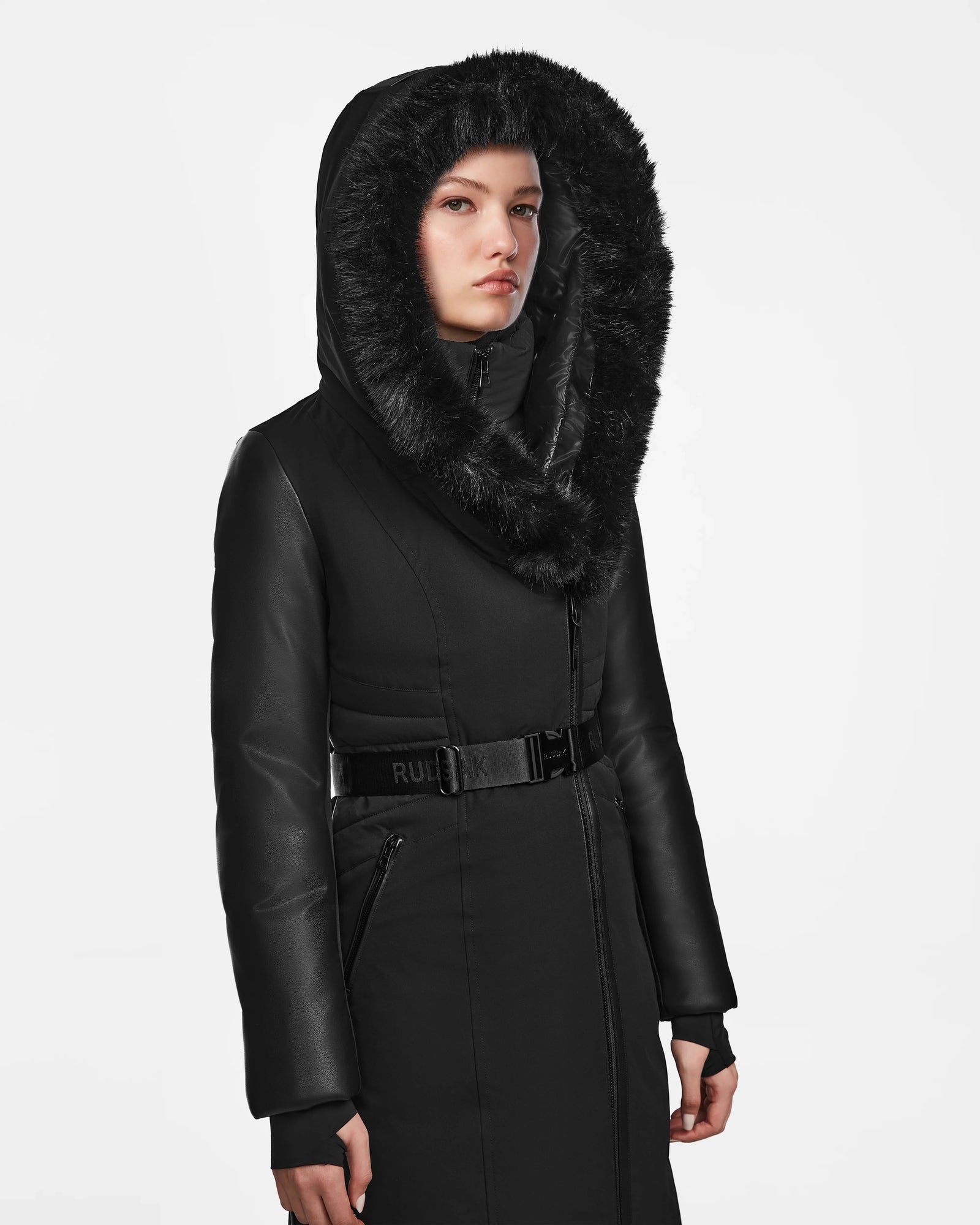 A woman models the Rudsak MISALA - L124512 Black womens long down coat with a fur-lined hood, belted waist, and leather-like sleeves. The water-resistant winter coat is showcased against a plain white background.