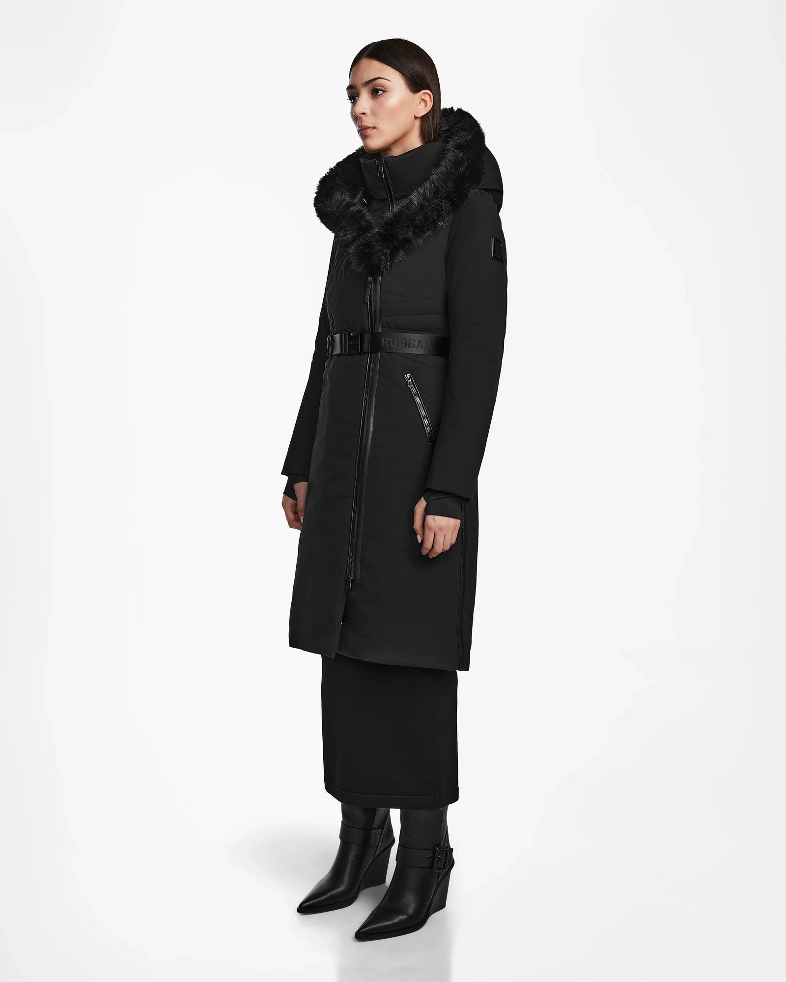 A woman stands in a studio wearing the Rudsak MUSA - L124534 Black women’s long down puffer with a fur-trimmed hood, belted waist, and zippered pockets. She pairs it with a black skirt and pointed ankle boots, facing slightly to the side.