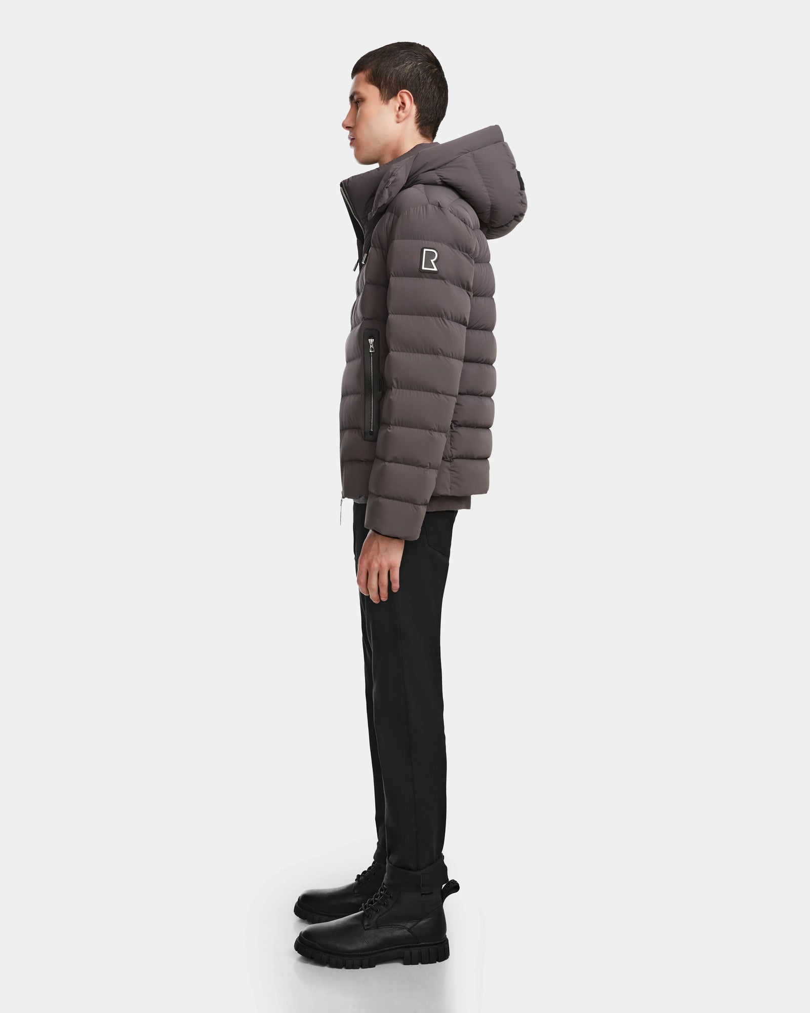 A person stands in profile wearing the Rudsak NAVAN - 6124551 Grey men’s quilted down puffer with a hood and “R” patch on the sleeve, paired with black pants and boots, against a plain white background.