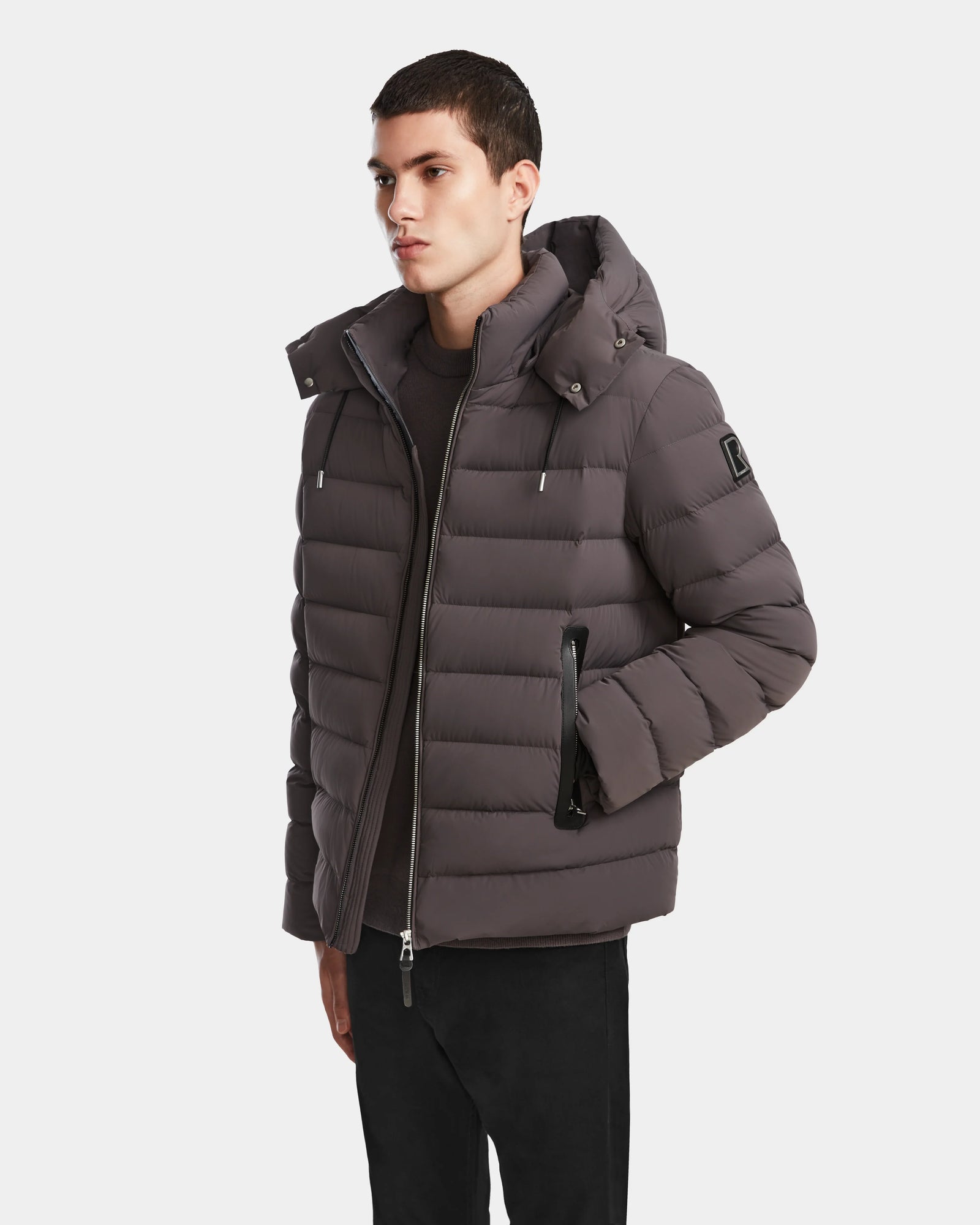 A person wearing the Rudsak NAVAN - 6124551 Grey mens down puffer jacket with a hood stands with one hand in their pocket against a plain light background.