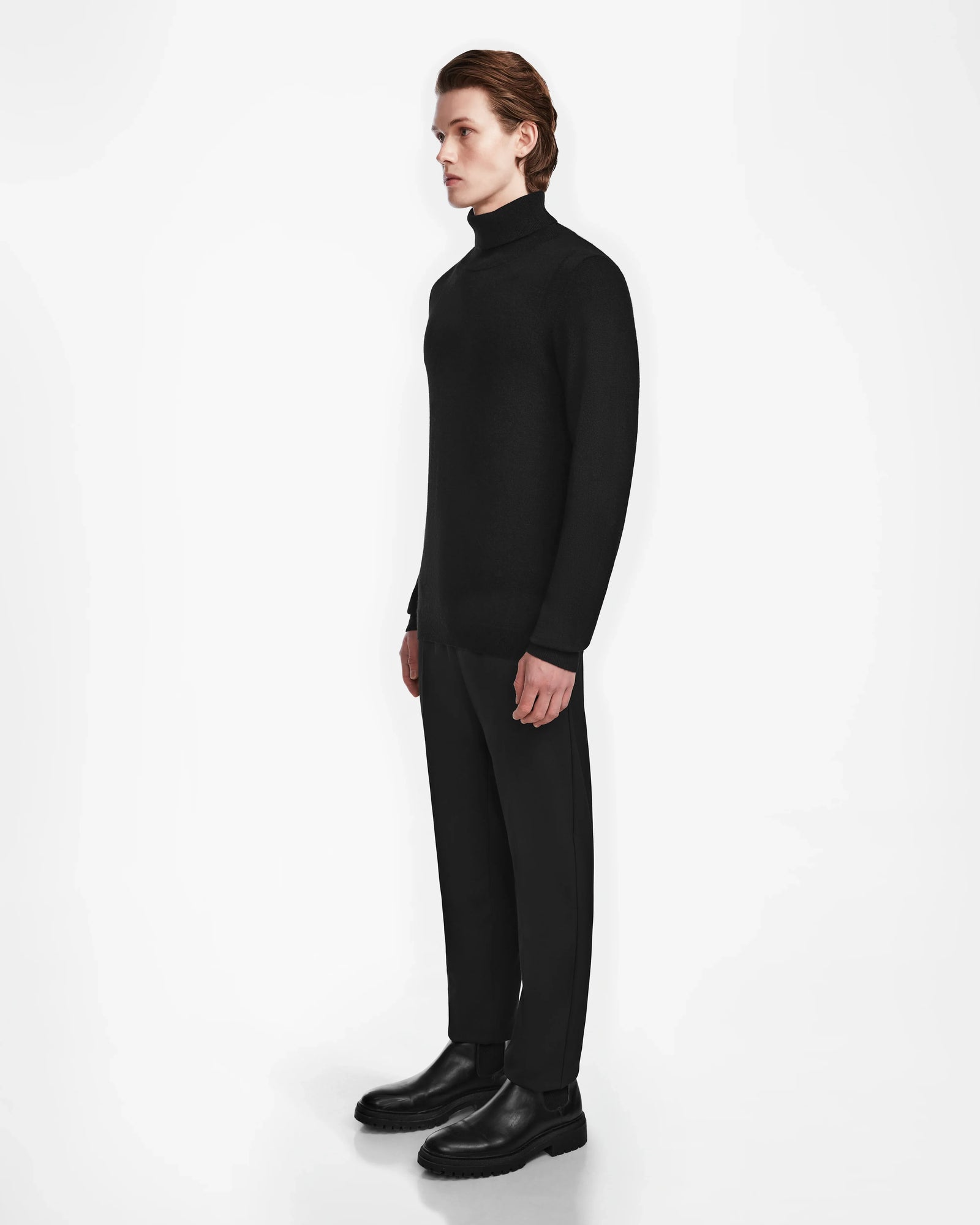 A person with light skin and straight, brown hair stands in profile against a white background, wearing the Rudsak NICOLAS - 6524528 Black mens cashmere turtleneck, along with black trousers and shoes.
