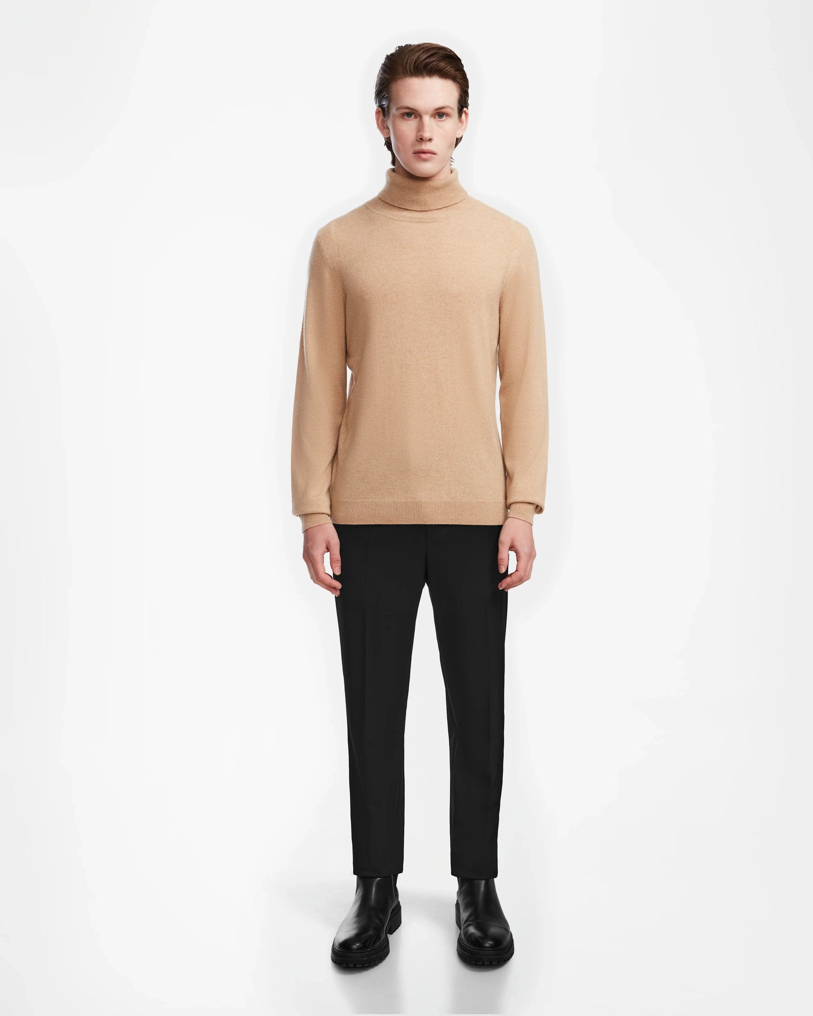 A young man faces forward wearing the Rudsak NICOLAS - 6524528 camel mens cashmere turtleneck, paired with black pants and black shoes, set against a plain white background.