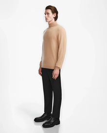 A person stands sideways against a white background, wearing the Rudsak NICOLAS - 6524528 Camel mens sweater, paired with black pants and black shoes.