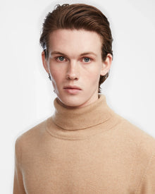 A young person with fair skin and short brown hair wears the Rudsak NICOLAS - 6524528 Camel mens luxury cashmere turtleneck sweater, looking directly at the camera against a plain white background.