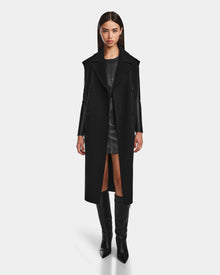 A woman with straight brown hair wears the Rudsak NOURA - 8124698 Black long coat over a wool vest, paired with a short black dress and knee-high boots, standing against a plain white background.