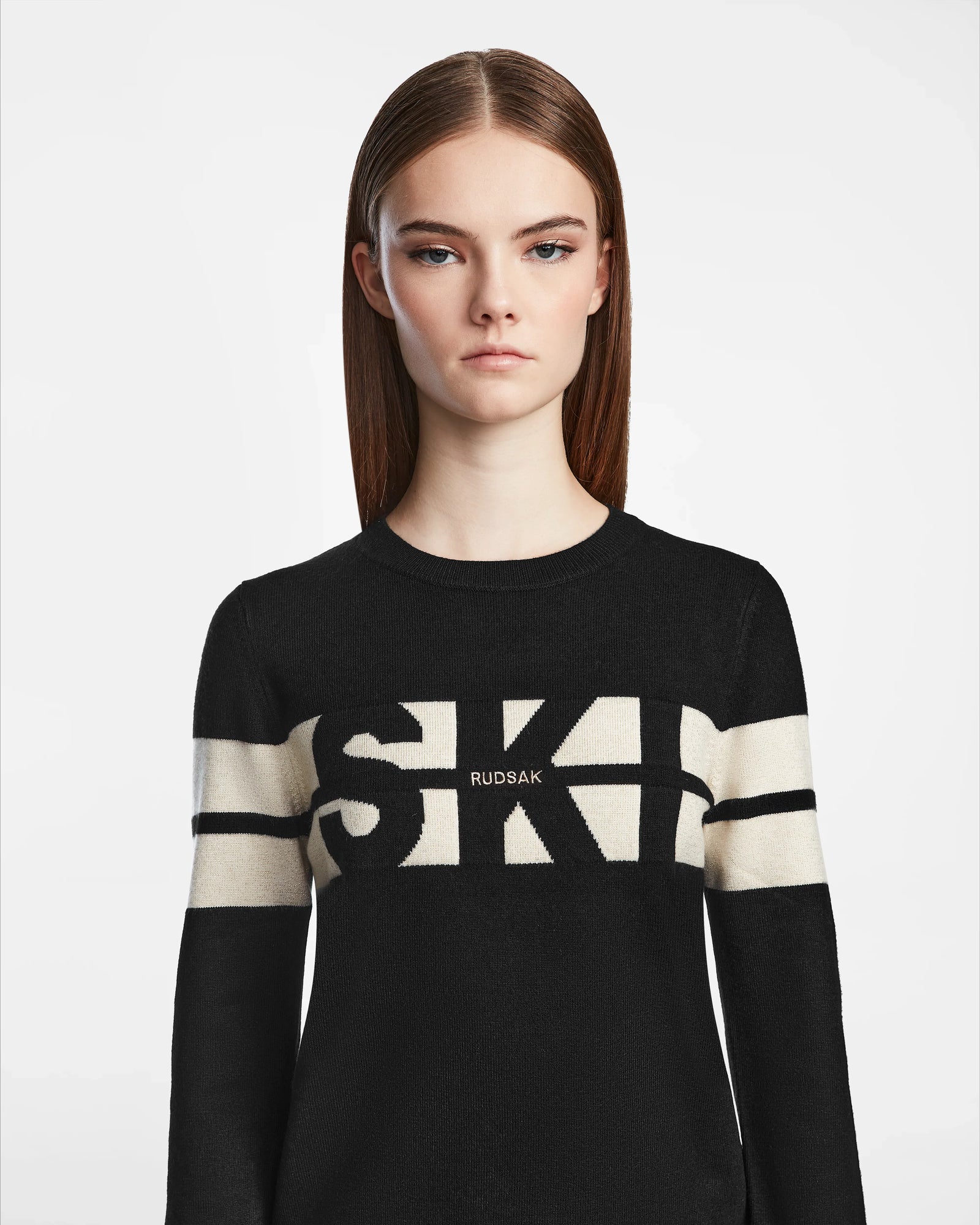 A woman with straight brown hair and a neutral expression wears the Rudsak ODESSA - 8523540 Black soft-knit sweater, featuring bold white SKI and RUDSAK lettering across the chest and arms. She stands against a plain white background.