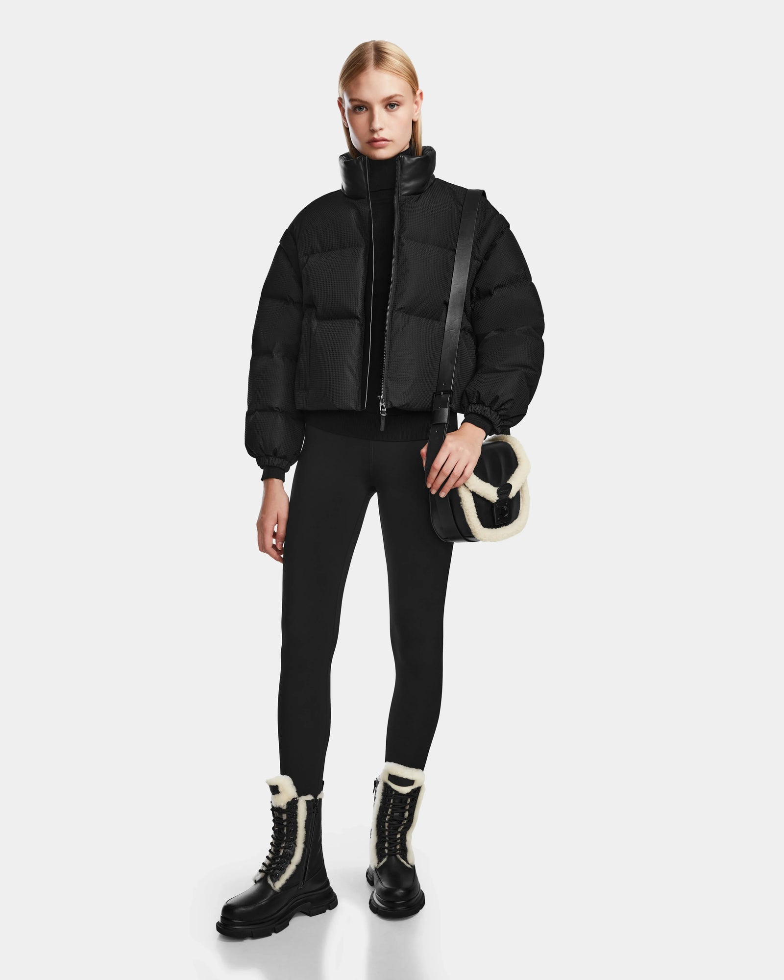 A woman wearing the Rudsak OPRAH - 8124708 Black cropped down puffer jacket, black leggings, black lace-up boots with white fur trim, and a black crossbody bag with white fur accents stands against a plain white background.