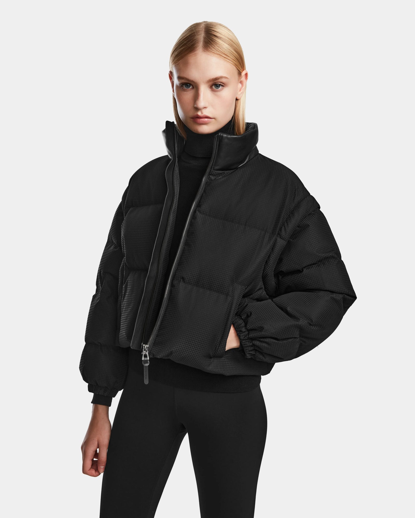 A woman with straight blonde hair models the Rudsak OPRAH - 8124708 Black womens cropped down puffer jacket with removable sleeves over a black turtleneck and leggings, standing against a plain light gray background.