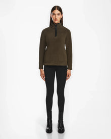 A woman faces forward against a plain white background wearing the Rudsak ORCHID - 8524616 Army dark olive fitted half-zip pullover sweater with vegan leather trims, paired with black leggings and black ankle boots.