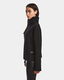 A woman with straight brown hair wears the Rudsak PARMI - 8524608 Black womens knit turtleneck sweater and black leather pants, standing in profile against a plain white background.