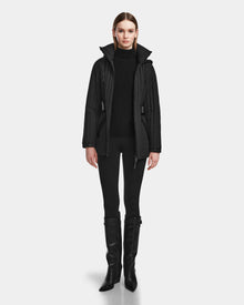 A woman faces forward in a Rudsak PETRAS - 8124725 Black vegan leather quilted jacket with a high collar, black turtleneck, fitted black pants, and knee-high black boots against a plain light background.
