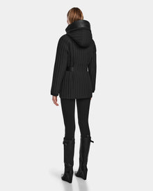 A person wearing the Rudsak PETRAS - 8124725 Black quilted jacket with a hood, black leggings, and knee-high boots stands with their back to the camera against a plain light gray background.