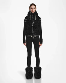 A person stands facing forward in the Rudsak RAQUEL S - 8124526 Black ski vest, black top, fitted pants, and large black fuzzy boots—ready for winter mountain adventures against a plain white background.