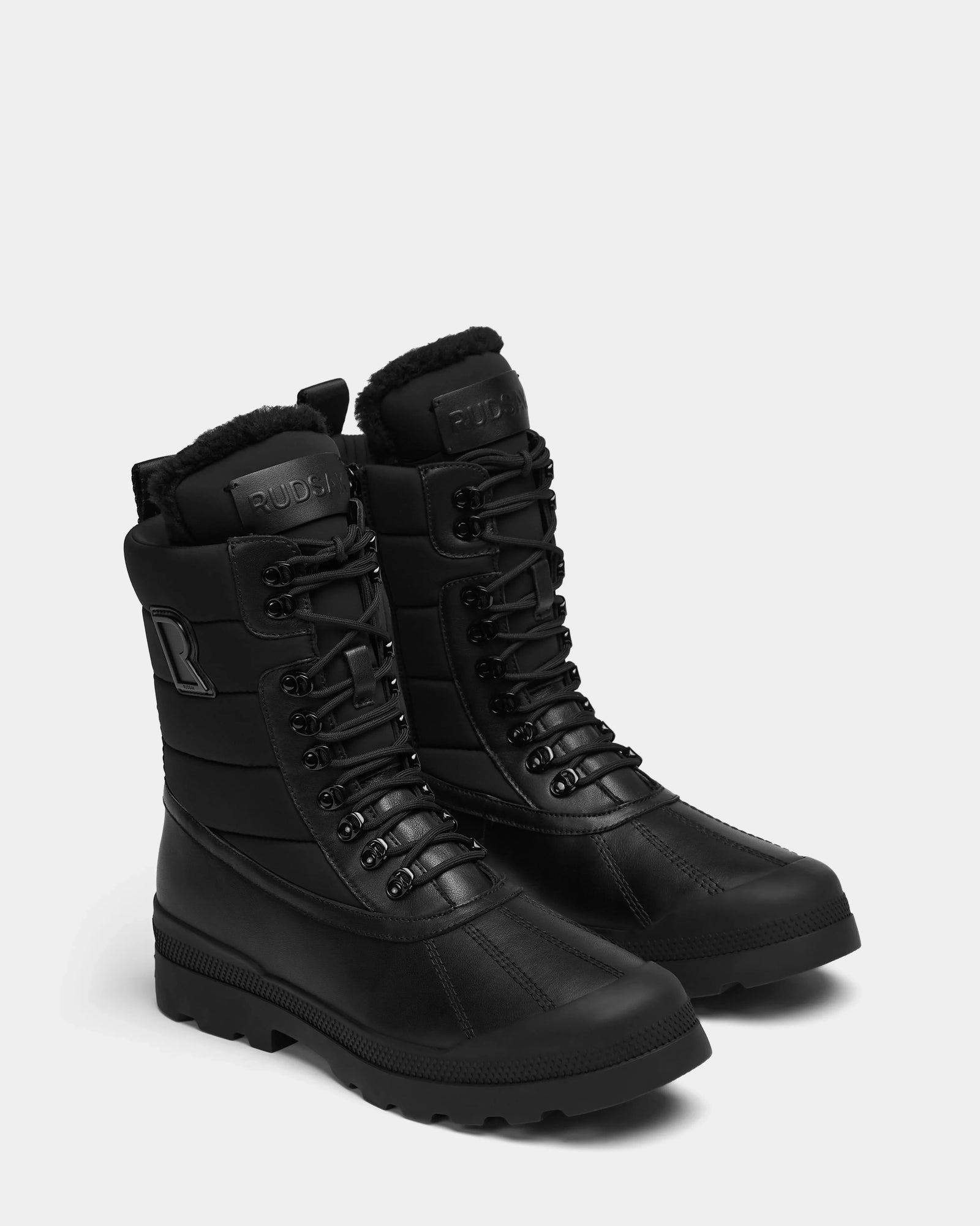 The Rudsak REAGAN - 6224501 Black men’s winter boots feature rugged soles, padded leather waterproof uppers, shearling lining, metal eyelets, and branding on the tongue and side, shown on a light gray background.