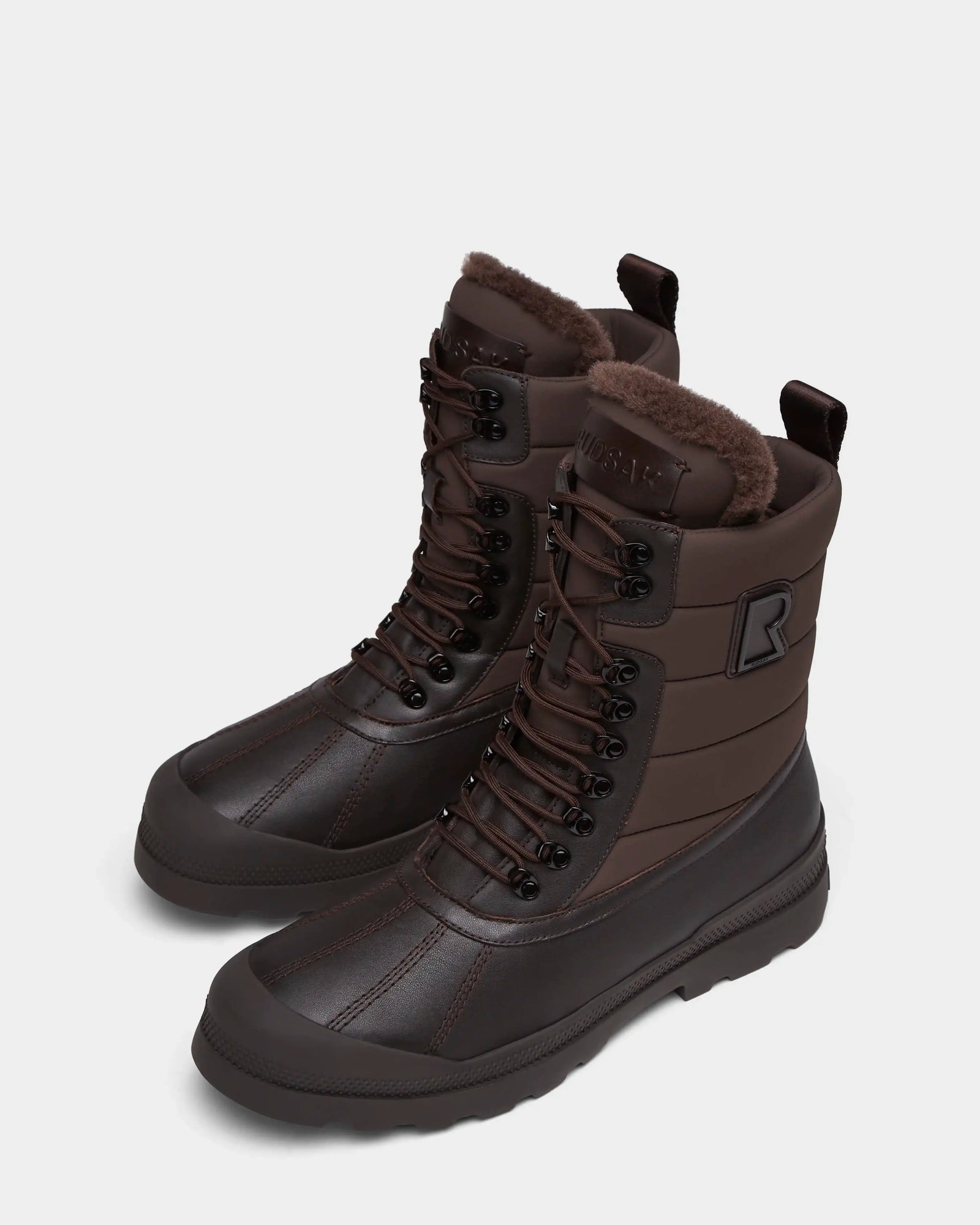 The REAGAN - 6224501 Espresso men’s winter boots by Rudsak feature dark brown leather, faux fur lining, black laces, rugged soles, a raised R logo on the sides, and sturdy pull tabs—perfect for cold weather.