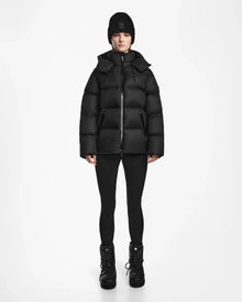 A person faces forward against a plain white background, wearing Rudsak's RITA M - L124537 Black weather-resistant puffer jacket, paired with black leggings, black winter boots, and a black knit beanie.