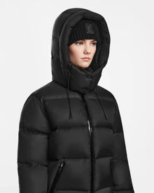 A person wears the Rudsak RITA M - L124537 Black women’s hooded down puffer with RDS-certified down, the hood up over a black beanie, looking forward with a neutral expression against a plain white background.