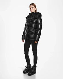 A woman with straight brown hair and a serious expression stands against a white background wearing the Rudsak RITA S - L124520 Black hooded down puffer, paired with black leggings and winter boots.