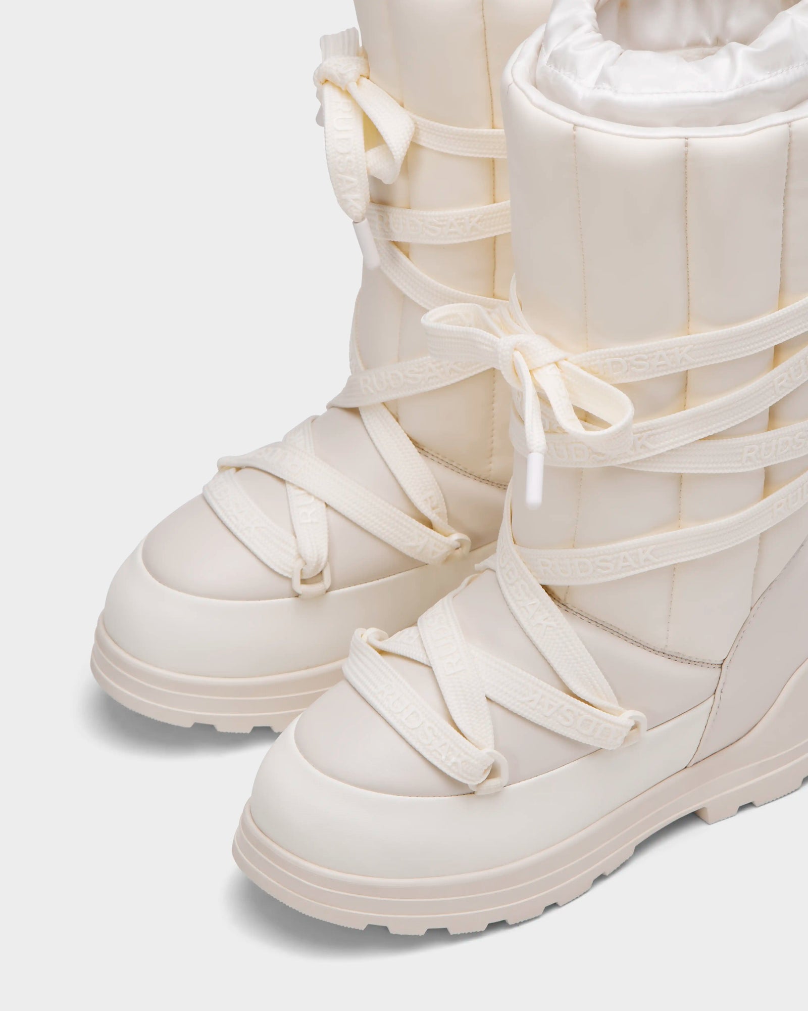 Close-up of the RODIN - 8224502 Iceland women’s winter boots by Rudsak, featuring off-white padded uppers, thick anti-slip soles, crisscross laces with “RUDSAK” branding, polar fleece lining for warmth, and a textured rubber sole for traction.