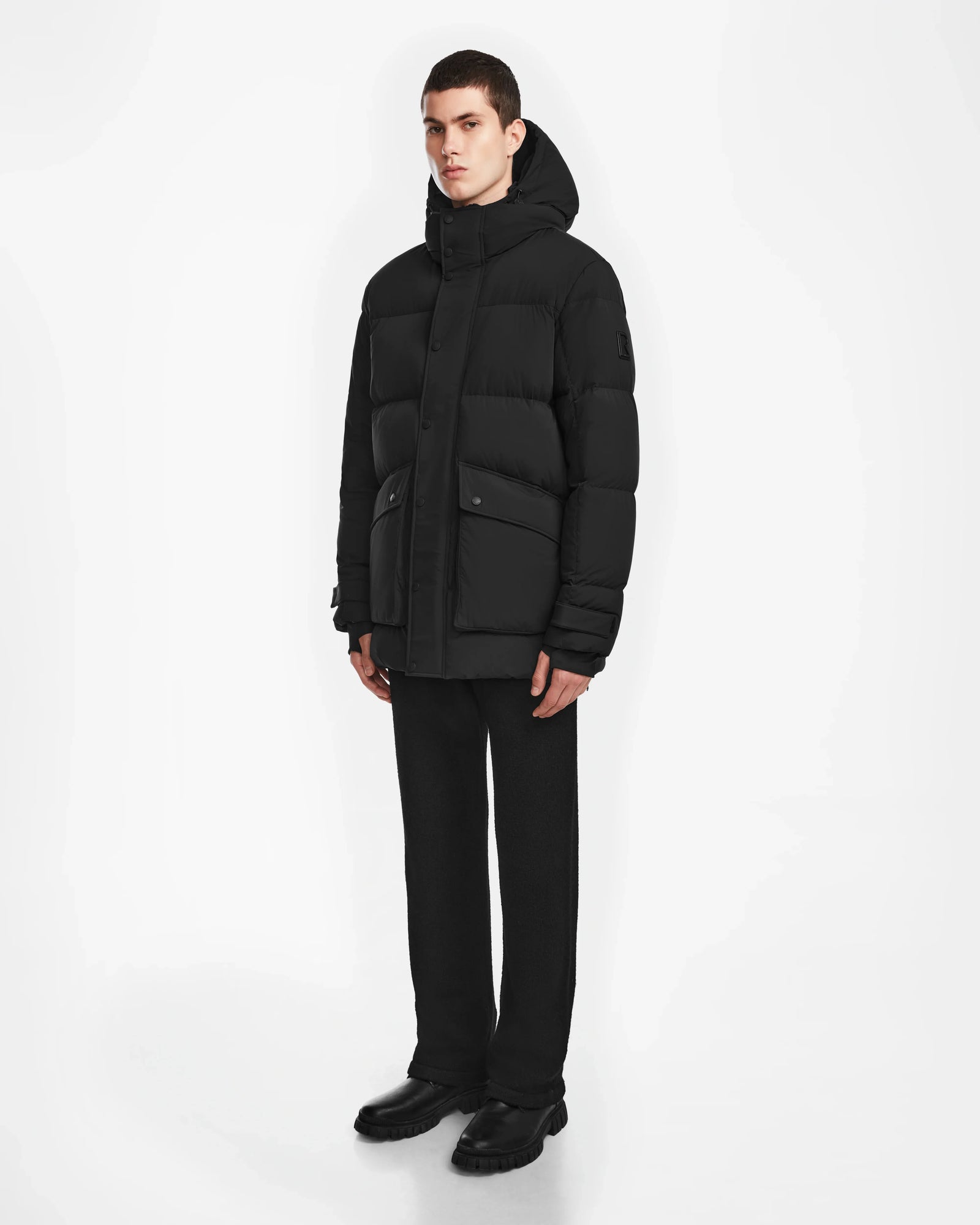 A man stands against a white background wearing the Rudsak ROKO - 6124510 Black quilted down parka, black pants, and black shoes. He faces forward with his hands at his sides, looking slightly to the left.
