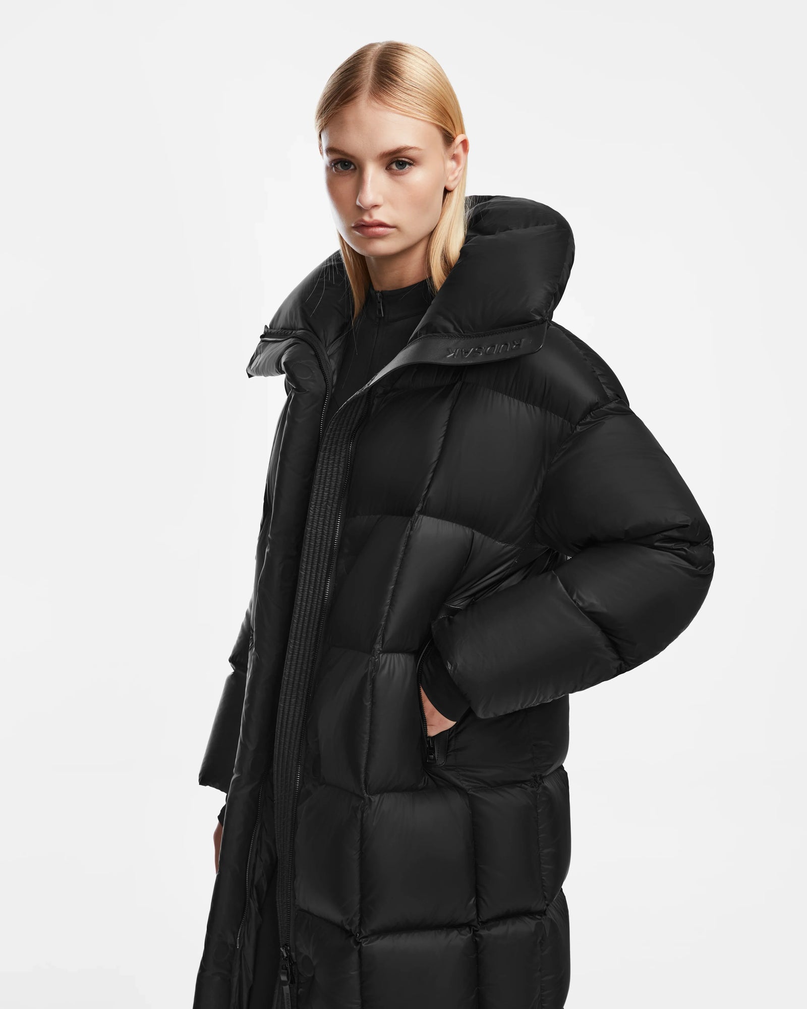 A woman with straight blonde hair wears the Rudsak SASH - 8124536 Black womens long down puffer coat, hands in pockets, standing against a plain white background and looking directly at the camera with a neutral expression.